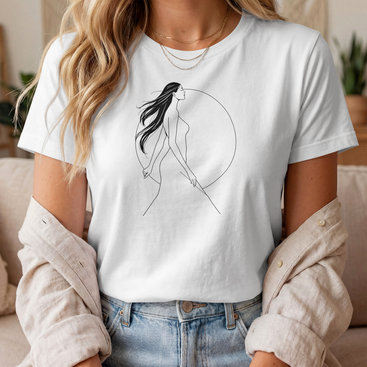A woman wearing a white short-sleeve crewneck t-shirt with a minimalist black line art graphic of a woman walking inside a circle. She has a beige button-down shirt off her shoulders and is wearing blue jeans.