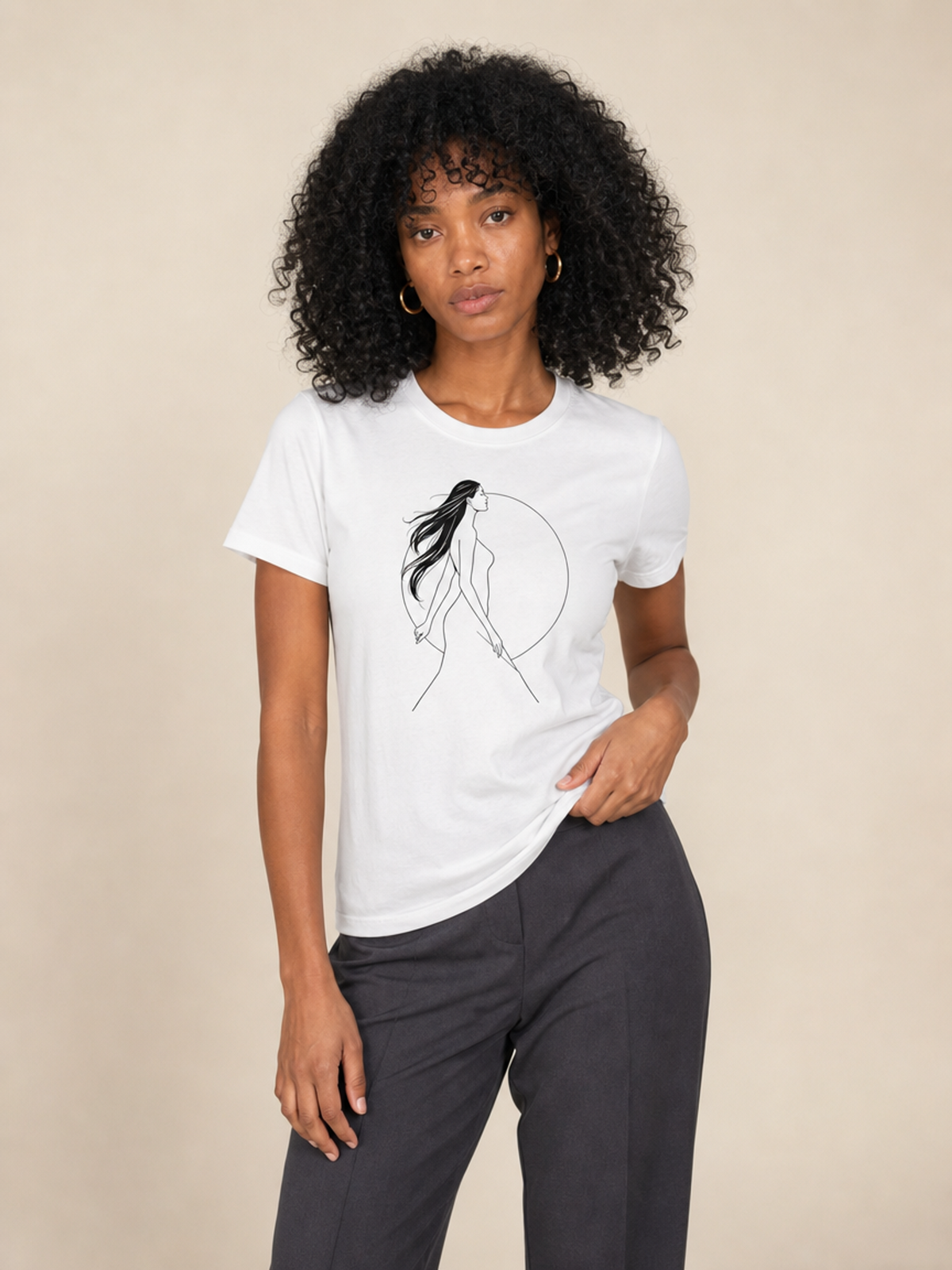 A woman with curly black hair wearing a white short-sleeve graphic t-shirt with a black line art illustration of a woman in a circle, paired with dark gray pants, against a solid beige background.