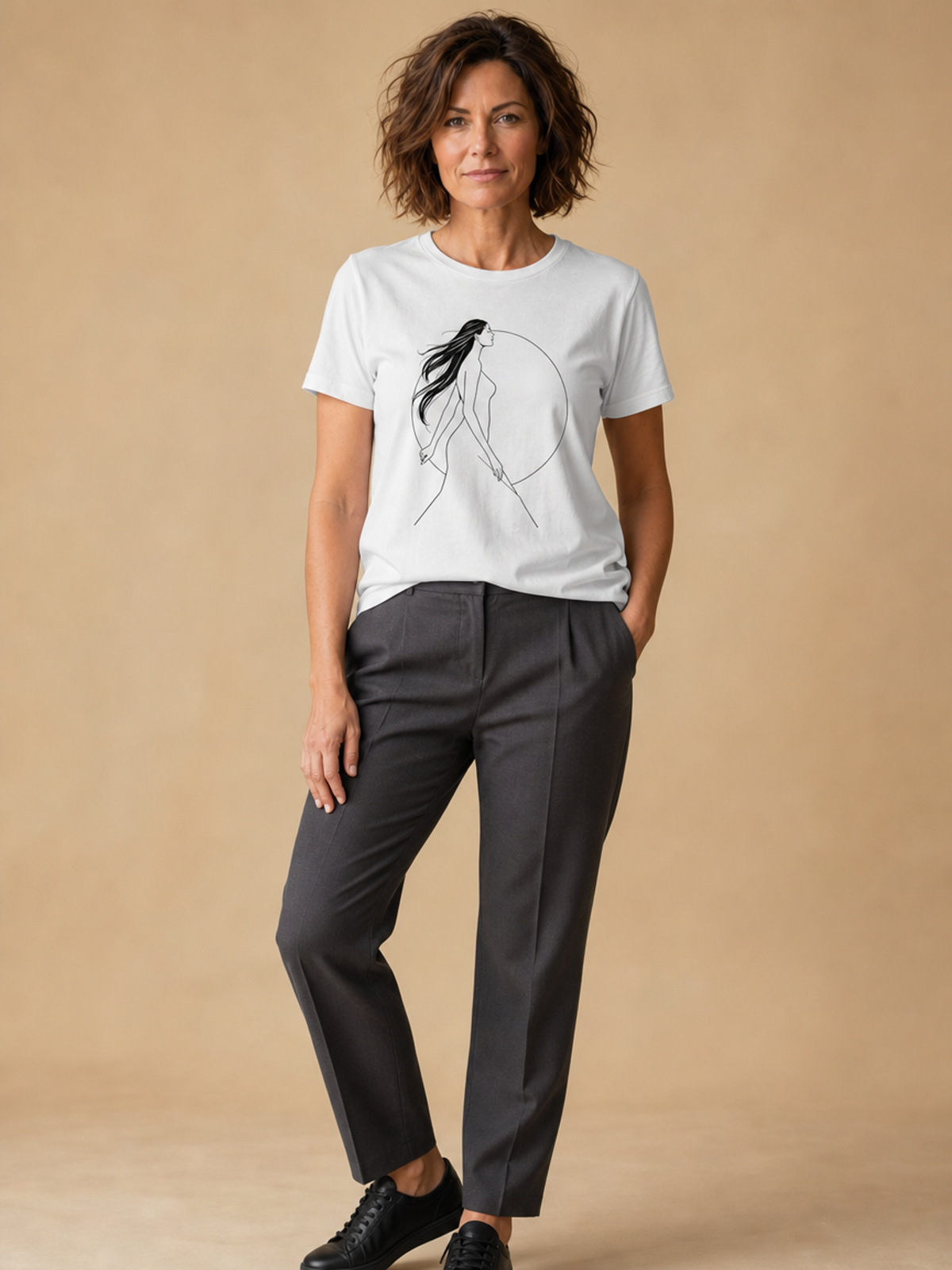 A woman modeling a white crewneck t-shirt with a minimalist black line art graphic of a woman in a circle, paired with dark gray trousers and black leather shoes, against a solid beige studio backdrop.
