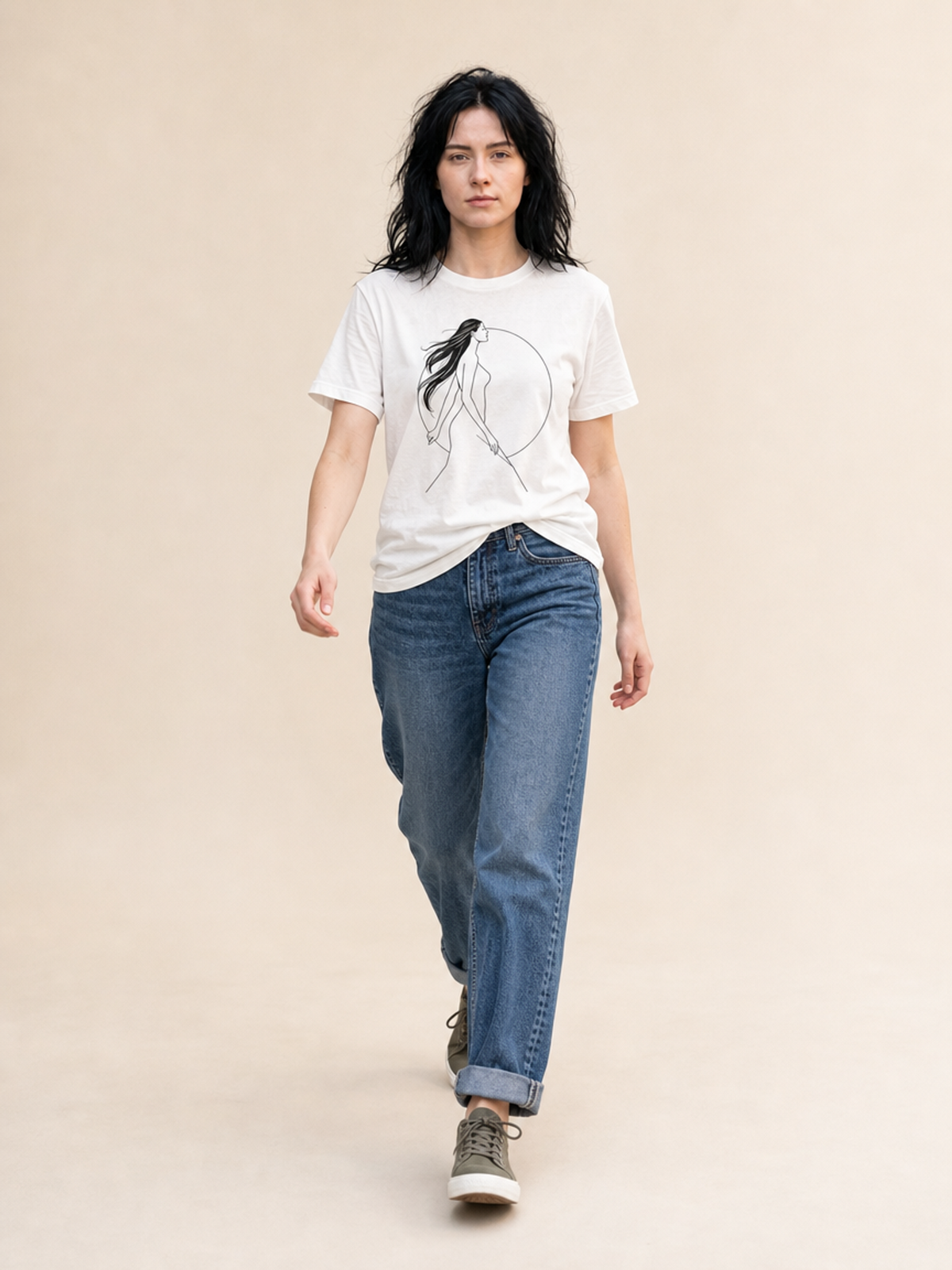 A model walks forward wearing a white graphic t-shirt with a black line art print of a woman, paired with blue jeans and gray sneakers, against a solid beige background.