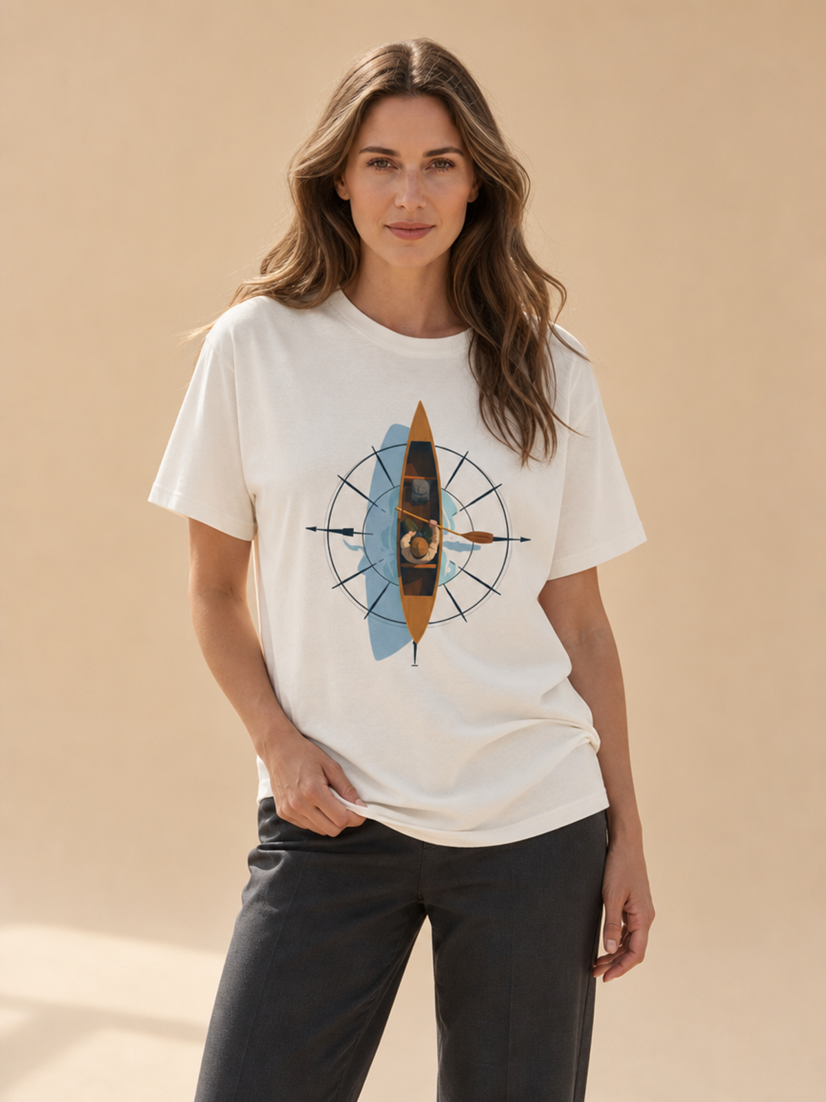 A woman modeling a white short-sleeve graphic T-shirt with a central compass design featuring a brown and gold kayak, paired with dark charcoal pants, against a warm beige studio background.