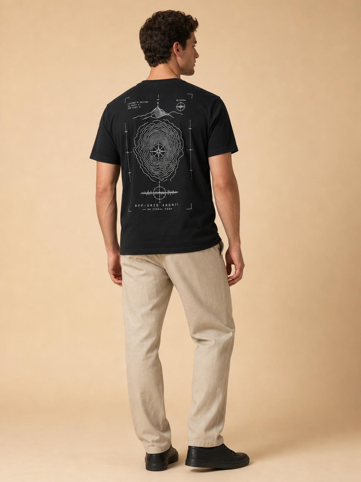 A man stands with his back to the camera wearing a black t-shirt with a large white technical blueprint-style graphic on the back, paired with beige pants and black shoes, against a solid beige studio backdrop.