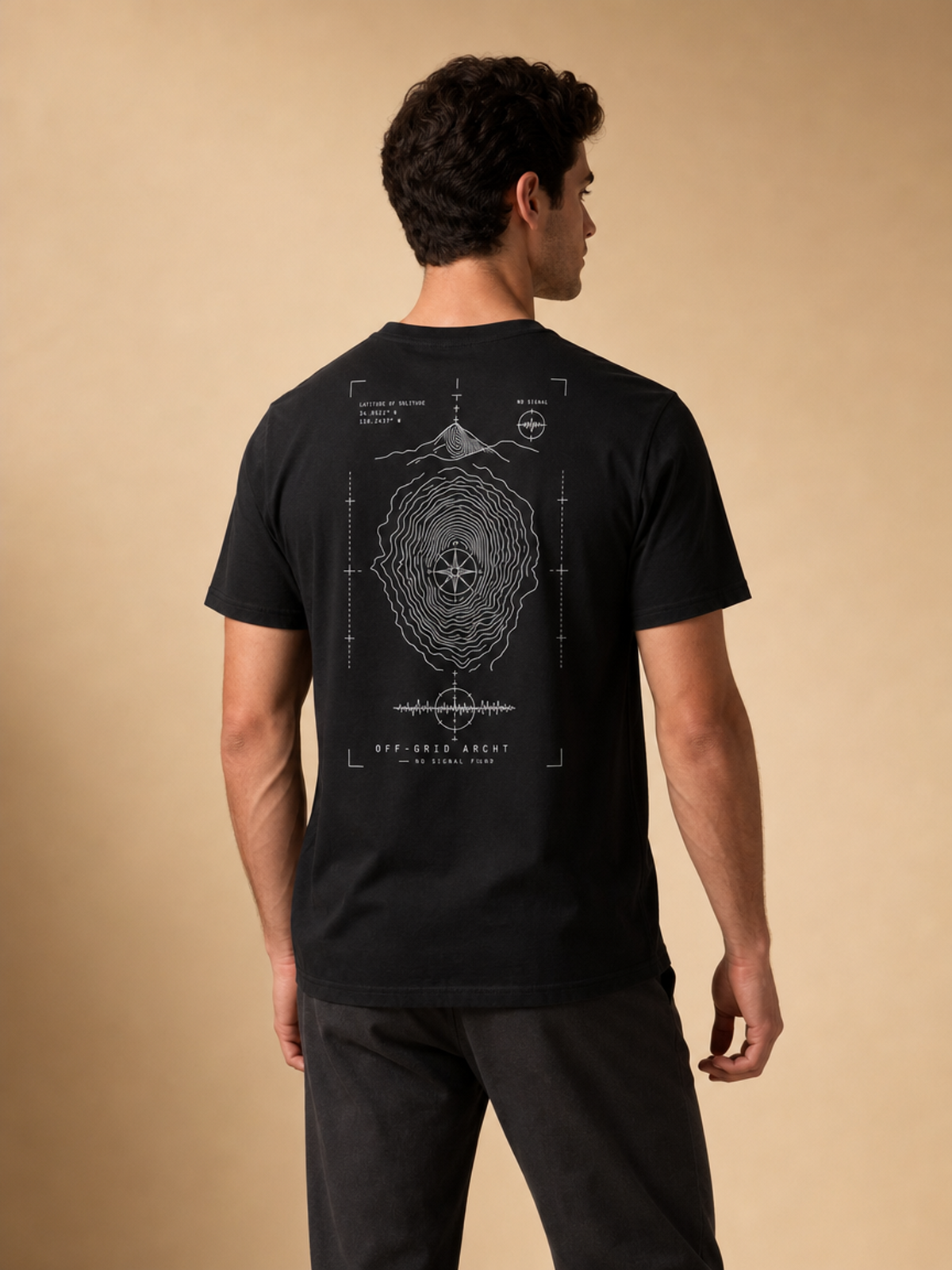 A man viewed from the back wearing a black t-shirt with a white technical blueprint-style graphic on the back, paired with dark gray pants, against a solid beige studio background.