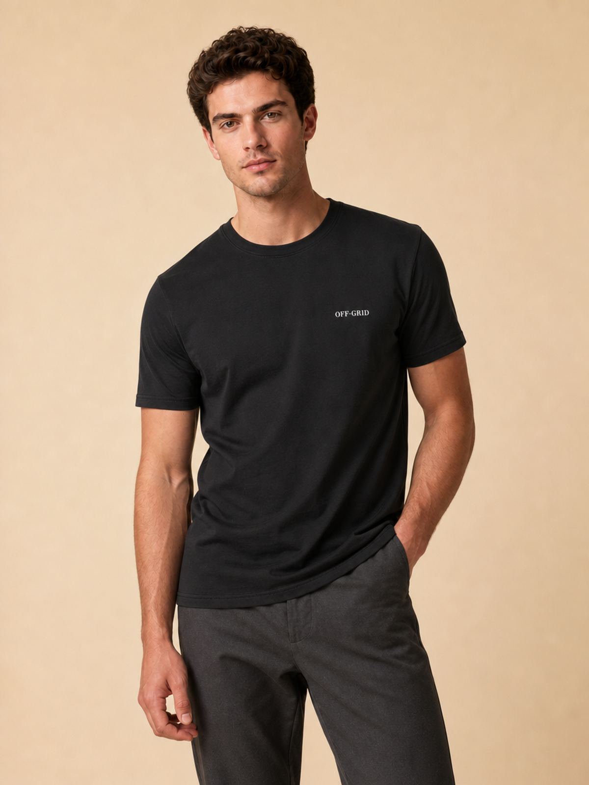 A man wearing a black short-sleeve crewneck t-shirt with 'OFF GRID' printed in white on the left chest, paired with dark gray pants, against a solid beige background.